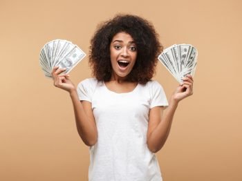 Portrait of a happy young african woman holding bunch of money banknotes isolated over beige background